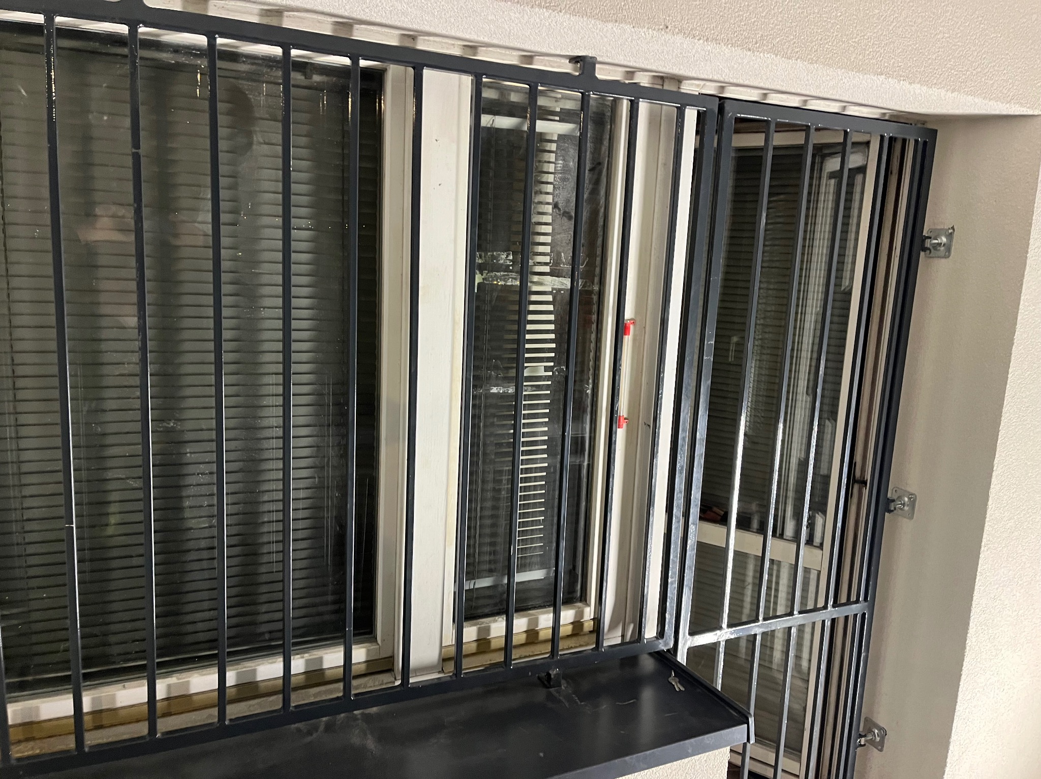 Zabezpieczenie okienne w Gliwicach: metalowa krata z uchylnym panelem, widok z zewnątrz, szare pręty na tle okna z żaluzjami.