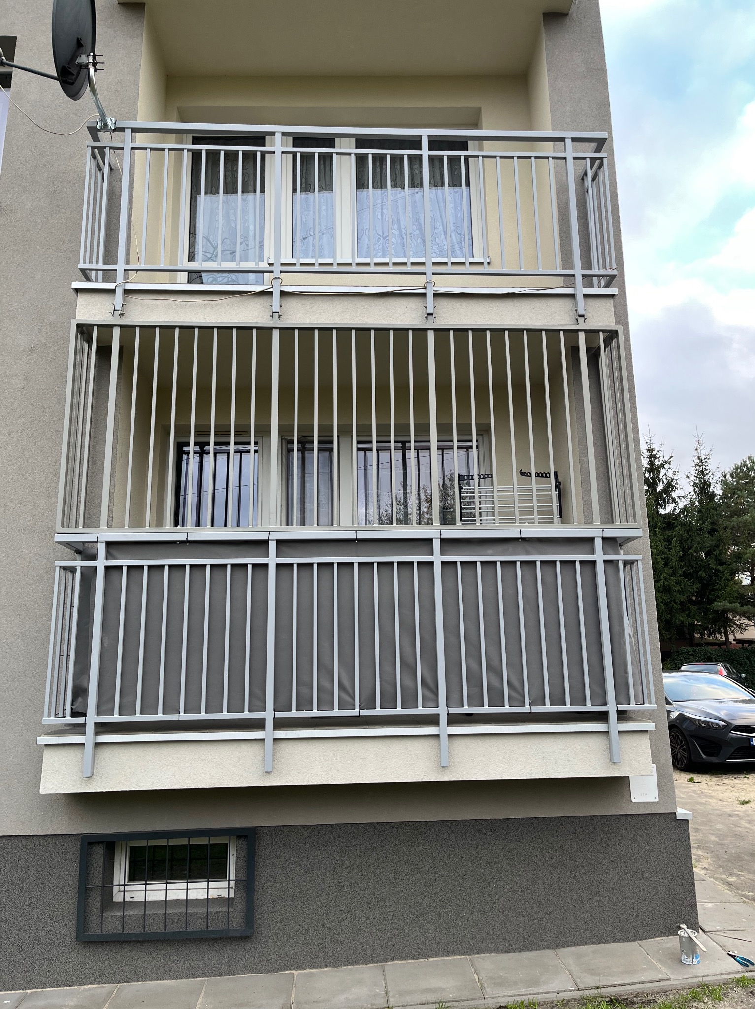 Trzy balkony jeden nad drugim z szarymi, metalowymi balustradami. Na najniższym balkonie dodatkowo szara tkanina. Na parterze okno z metalową kratą.