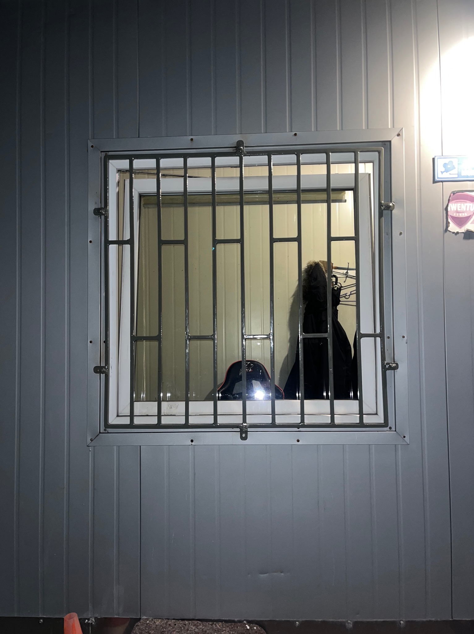 Kraty okienne z metalowymi prętami na szarym budynku z blachy trapezowej, widoczne ubrania w środku. Zabezpieczenie okna.