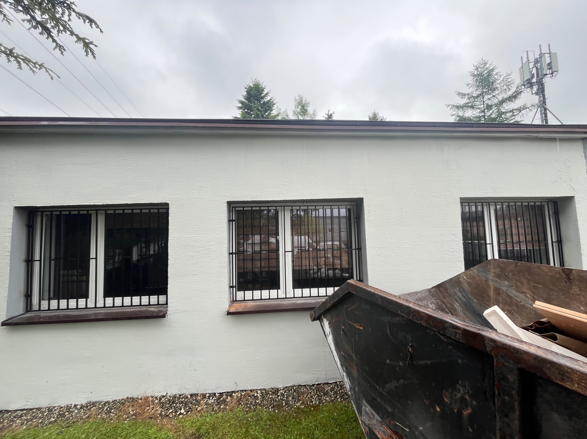 Okna z kratami ochronnymi w Gliwicach, widoczne z zewnątrz budynku. Kontener na gruz w prawym dolnym rogu. Szare niebo w tle.