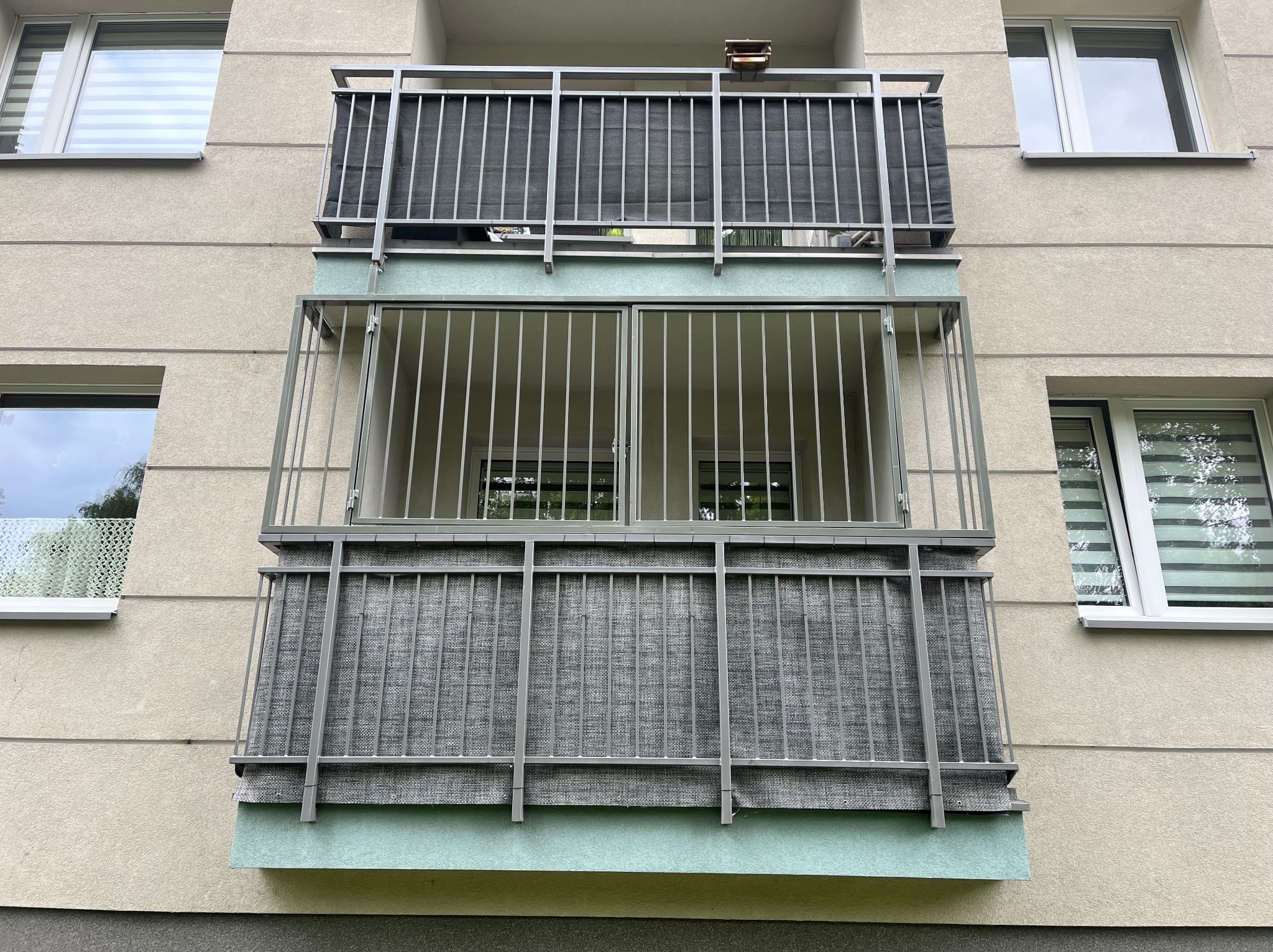 Dwa balkony z metalowymi balustradami i szarą osłoną, zamontowane na elewacji budynku z oknami. Górny balkon z budką dla ptaków.
