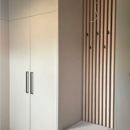 FIX&CARE SŁAWOMIR KOSNO - Biała szafa wnękowa z czarnymi uchwytami i drewnianym panelem z wieszakami, uzupełniona siedziskiem z szarej tapicerki. Nowoczesny design.