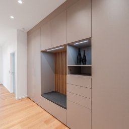 FIX&CARE SŁAWOMIR KOSNO - Beżowa szafa wnękowa z siedziskiem i półkami, oświetlenie LED, drewniane lamele, podłoga z jasnego drewna, minimalistyczny design, nowoczesne wykończenie.