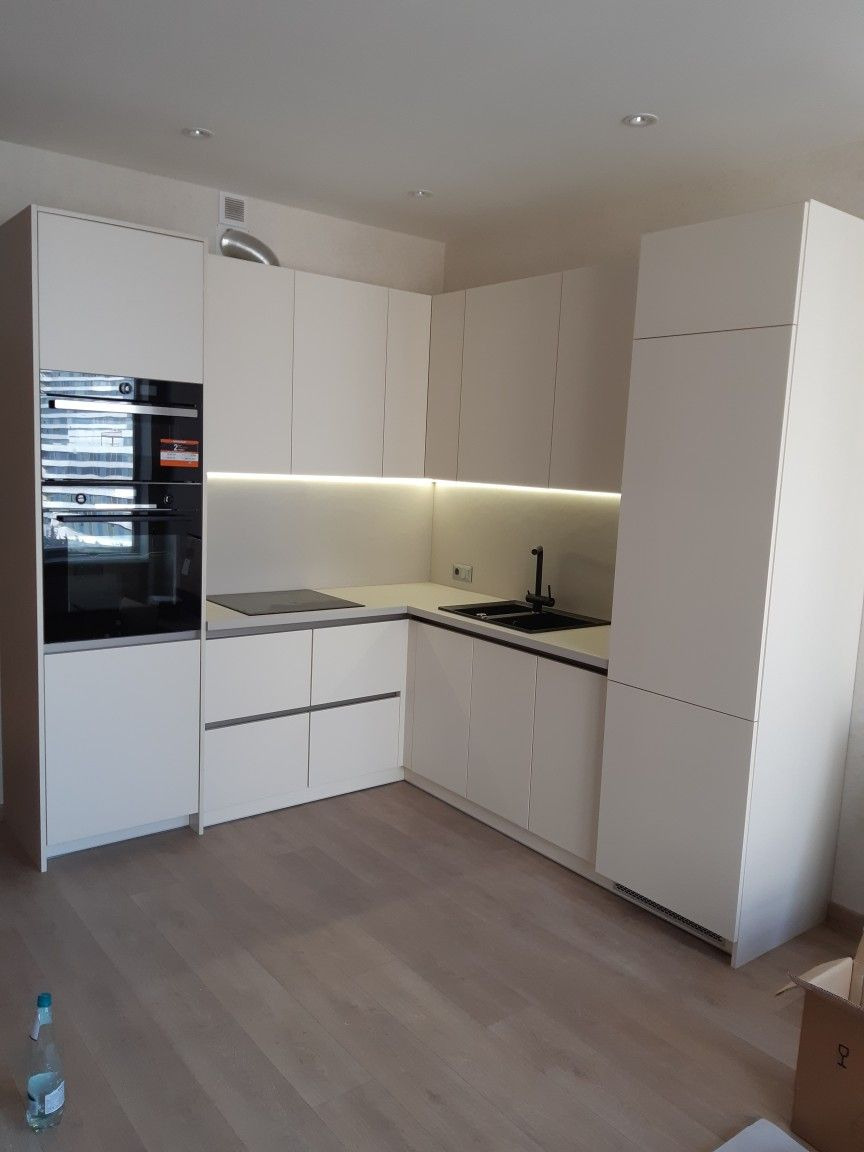 Nowoczesna, minimalistyczna kuchnia z białymi szafkami i czarną armaturą. Podświetlenie LED pod szafkami górnymi. Jasna podłoga z paneli. Widok na całe pomieszczenie.