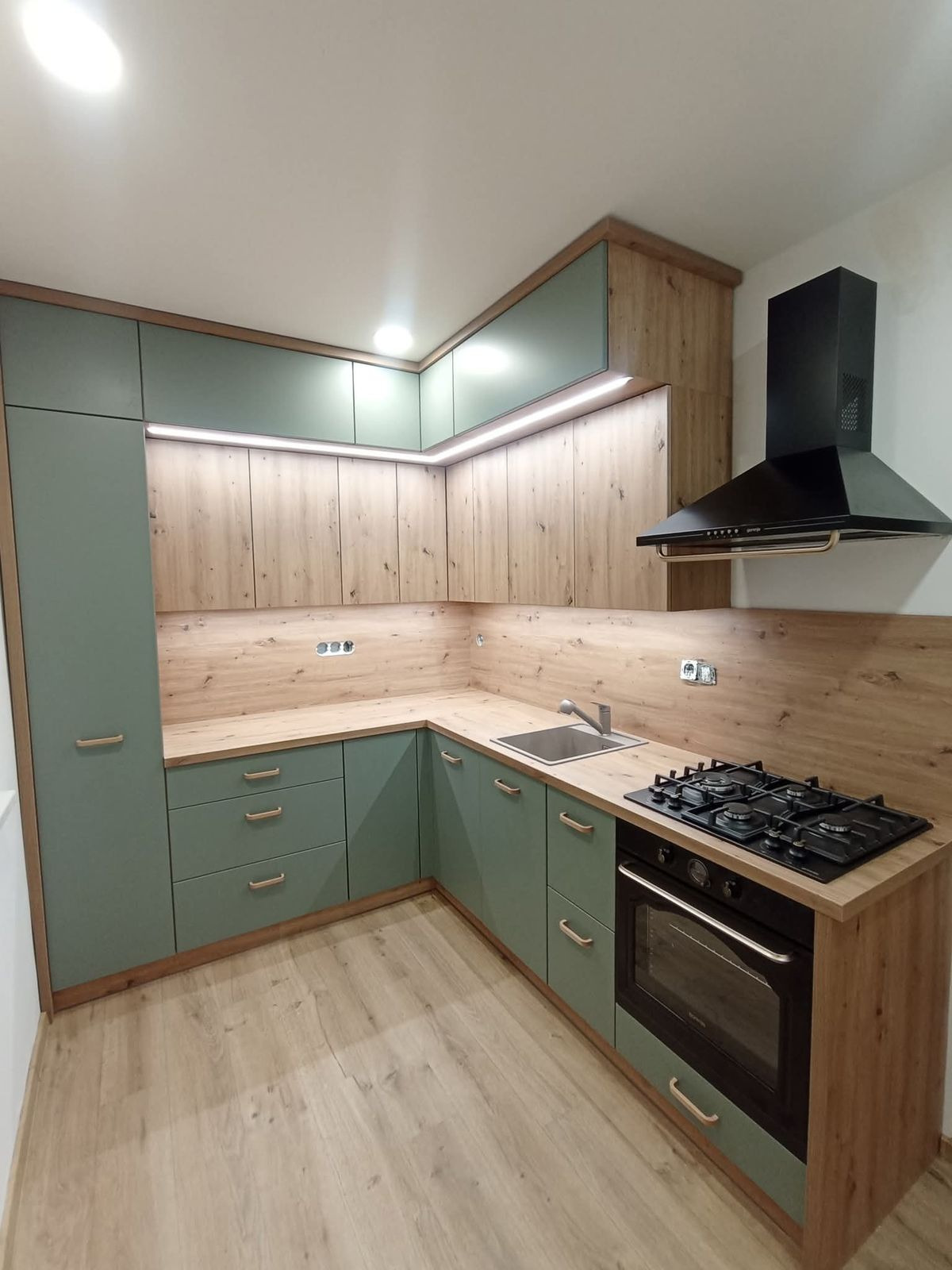 Nowoczesna kuchnia w odcieniach zieleni i drewna z oświetleniem LED podszafkowym, czarnym okapem i płytą gazową. Ujęcie z perspektywy narożnika.