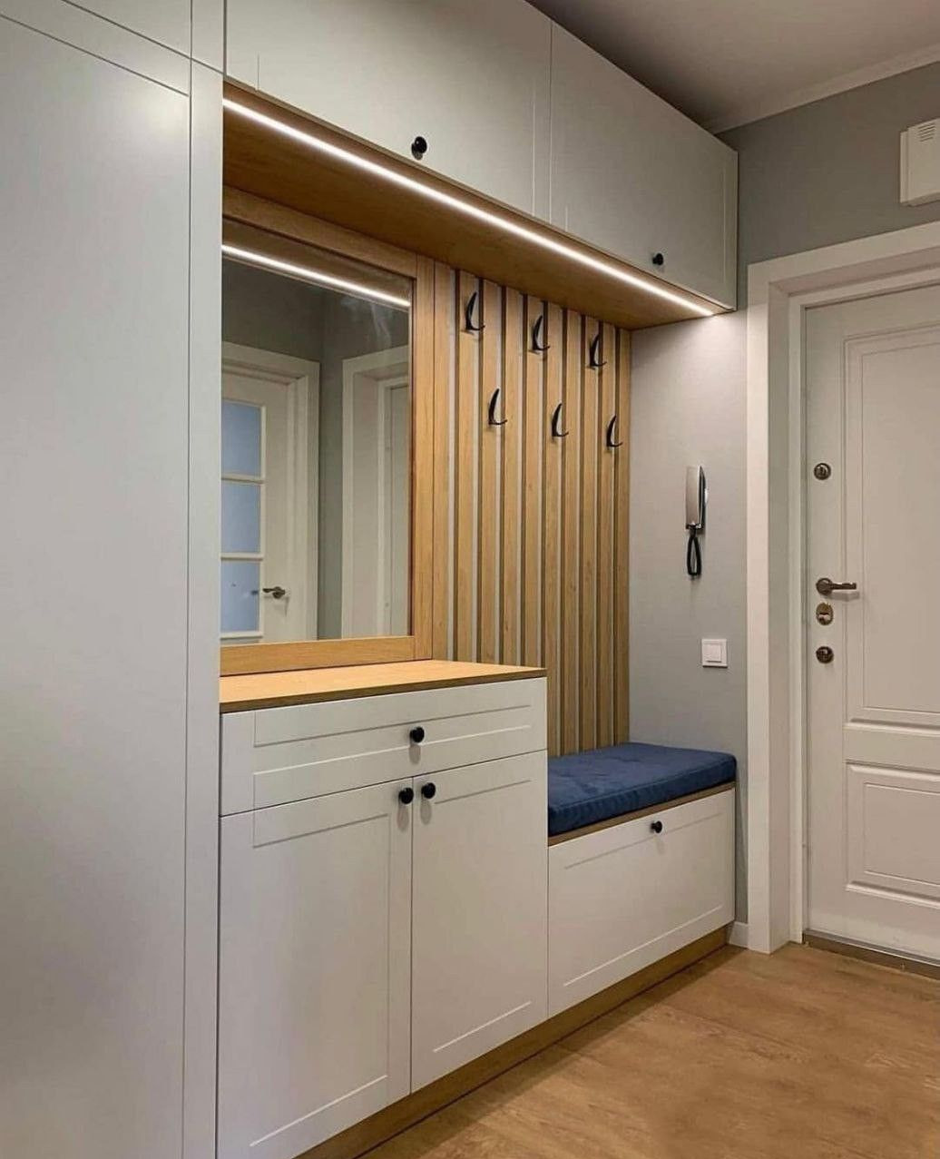 Funkcjonalna zabudowa przedpokoju: białe szafki z siedziskiem, lustro i drewniany panel z haczykami, oświetlenie LED. Nowoczesny design, jasne kolory.