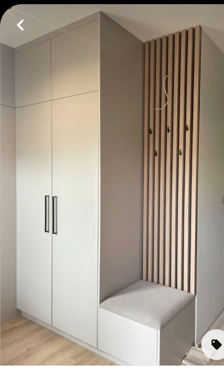 Biała szafa wnękowa z czarnymi uchwytami i drewnianym panelem z wieszakami, uzupełniona siedziskiem z szarej tapicerki. Nowoczesny design.