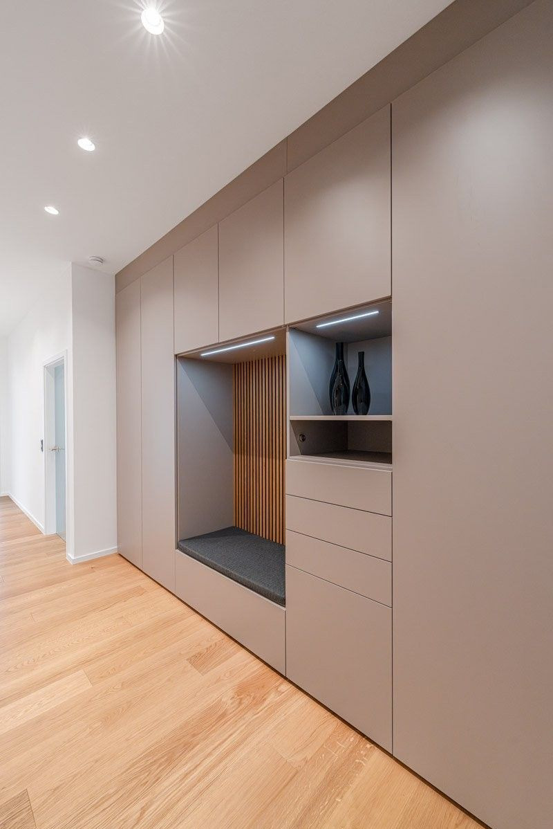 Beżowa szafa wnękowa z siedziskiem i półkami, oświetlenie LED, drewniane lamele, podłoga z jasnego drewna, minimalistyczny design, nowoczesne wykończenie.