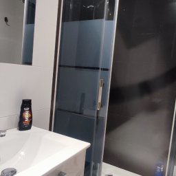 TOMNOR - Nowoczesna łazienka z kabiną prysznicową z chromowanymi profilami, białą umywalką z szafką i czarnymi płytkami na ścianie.