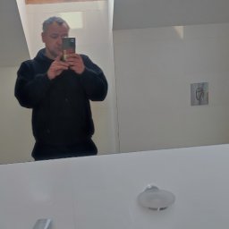 TOMNOR - Mężczyzna robiący sobie zdjęcie w lustrze w nowocześnie wykończonej łazience z oknem dachowym, widoczna biała umywalka, bateria i dozownik na mydło.