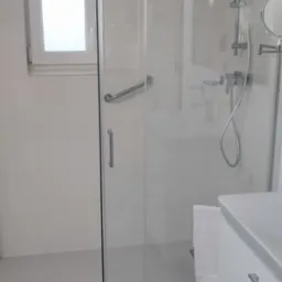 Nowoczesna, jasna łazienka z kabiną prysznicową ze szklanymi przesuwnymi drzwiami, chromowaną armaturą, umywalką z szafką z szufladami oraz małym oknem.