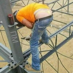TOMNOR - Spawacz w pomarańczowej koszulce i dżinsach spawa stalową konstrukcję na zewnątrz, widoczny fragment bielizny ponad spodniami.