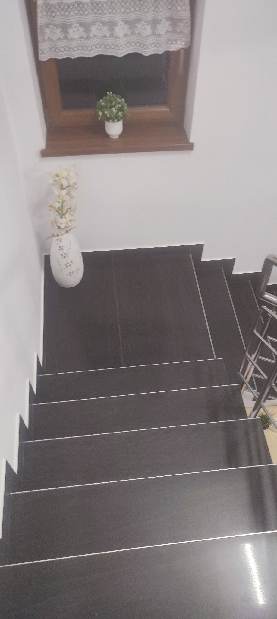 Schody wyłożone ciemnymi, błyszczącymi płytkami, z białymi fugami, widziane z góry, z dekoracjami w postaci kwiatów w wazonie i doniczce na parapecie