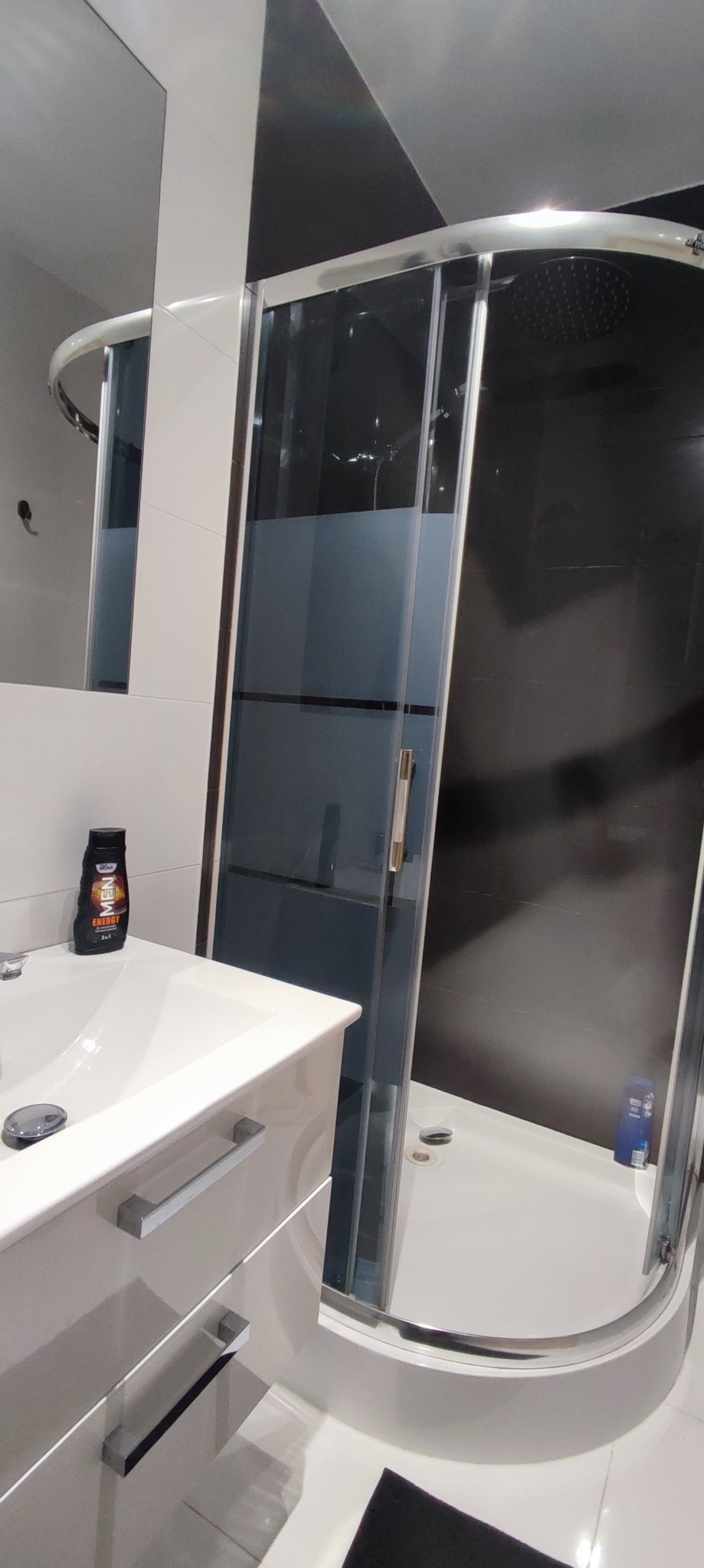 Nowoczesna łazienka z kabiną prysznicową z chromowanymi profilami, białą umywalką z szafką i czarnymi płytkami na ścianie.