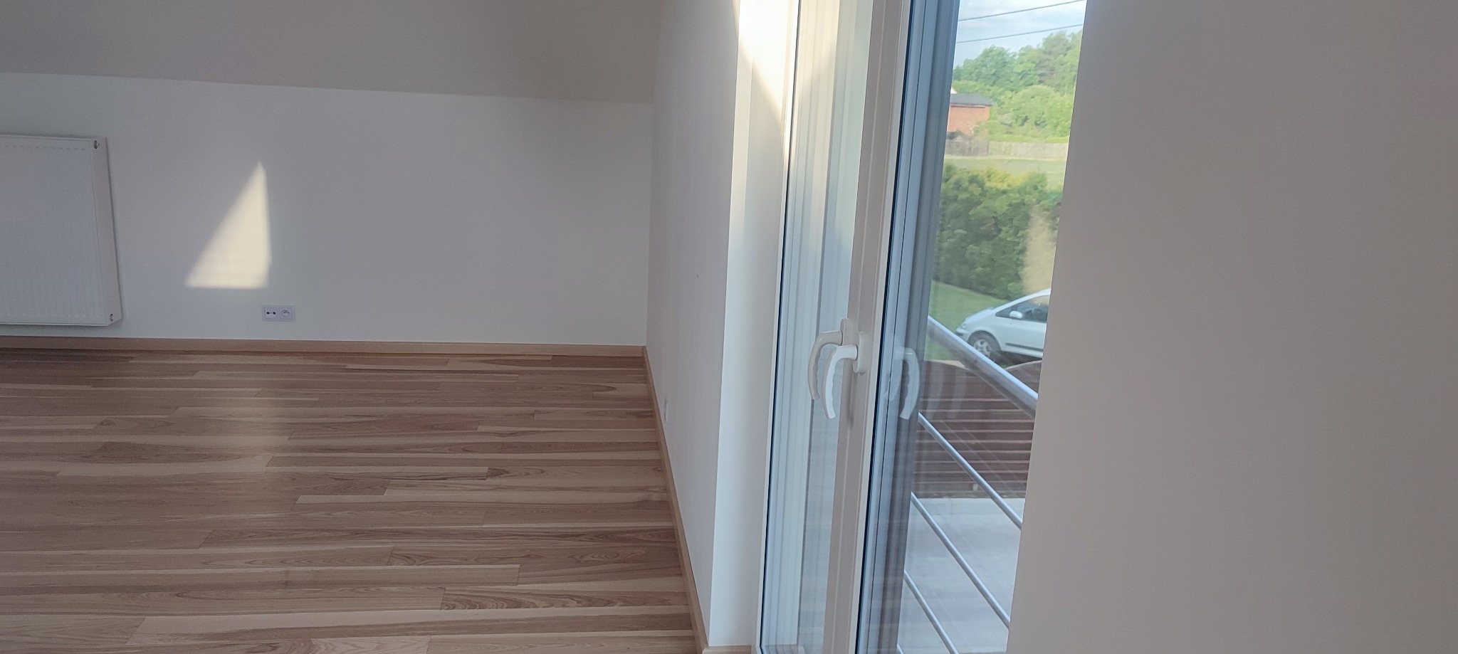 Jasne, puste poddasze z parkietem i widokiem na zewnątrz przez okno balkonowe, widoczny fragment grzejnika i gniazdka elektrycznego.