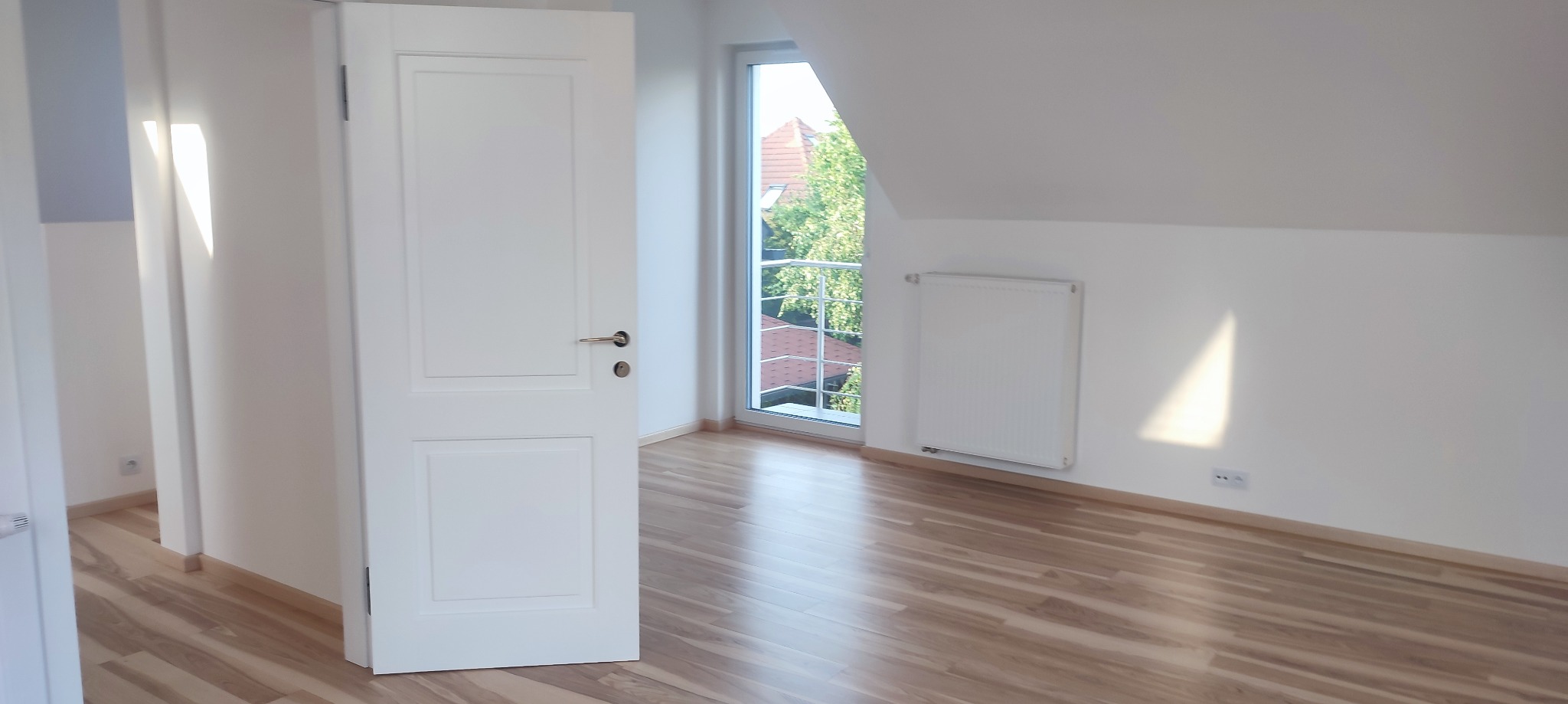 Jasne, puste poddasze z drewnianą podłogą, białymi ścianami, oknem z widokiem na zieleń i białym grzejnikiem ściennym. Białe drzwi wewnętrzne z srebrną klamką są otwarte, wpadające światło...