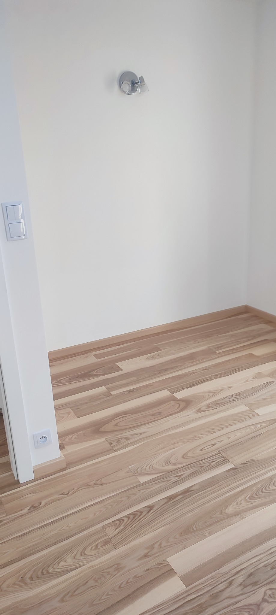 Jasne panele podłogowe ułożone diagonalnie w rogu pokoju z białymi ścianami, widoczny kontakt elektryczny i chromowana lampa sufitowa.