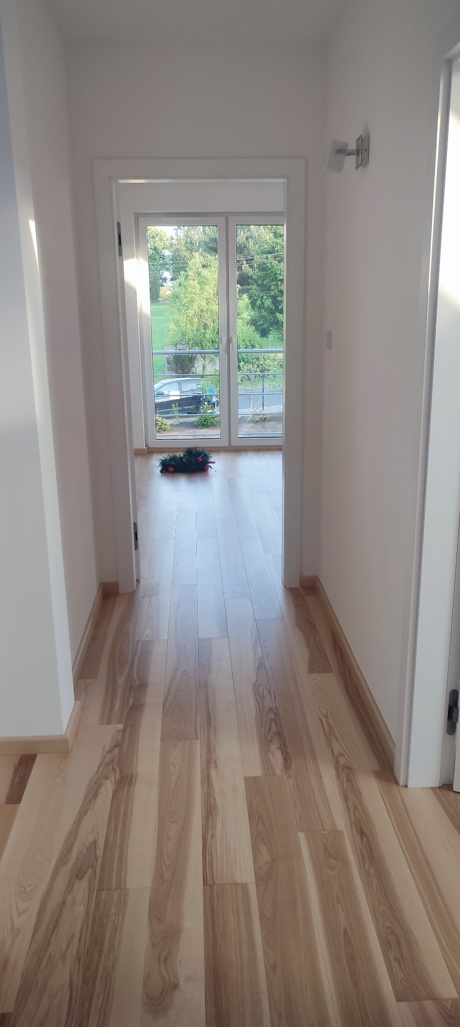 Korytarz z jasną, drewnianą podłogą ułożoną w deski, białe ściany i framugi drzwi, z widokiem na balkon i zieleń za oknem. Na podłodze leży świąteczny wieniec.