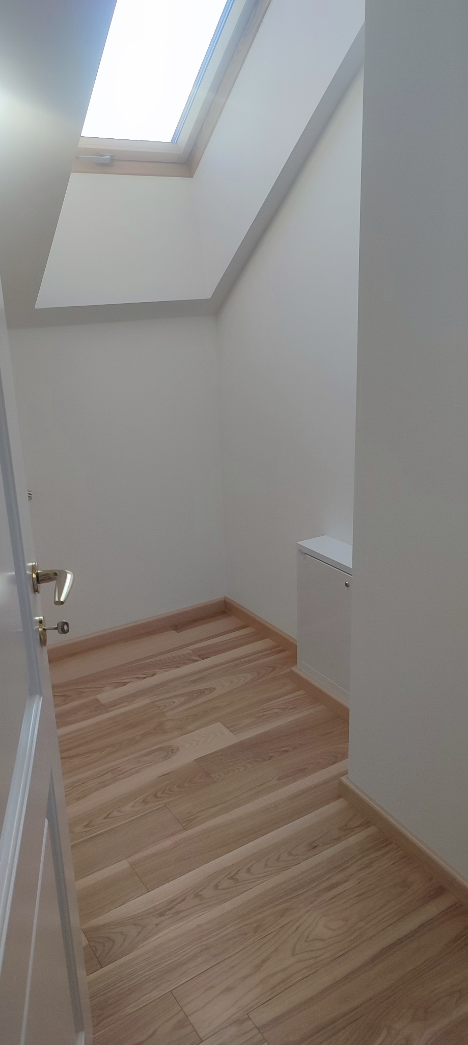 Jasne drewniane panele podłogowe ułożone ukośnie w małym pomieszczeniu z oknem dachowym i białą szafką w rogu, widok zza uchylonych drzwi.