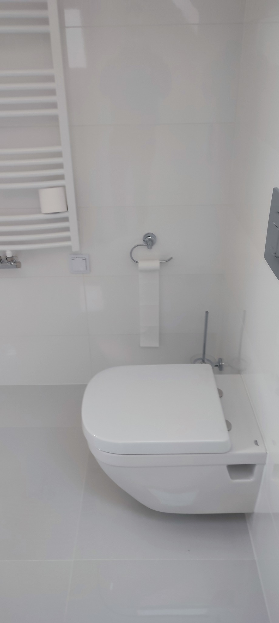 Nowoczesna, minimalistyczna toaleta podwieszana w łazience wykończonej białymi płytkami, z grzejnikiem drabinkowym i uchwytem na papier toaletowy.