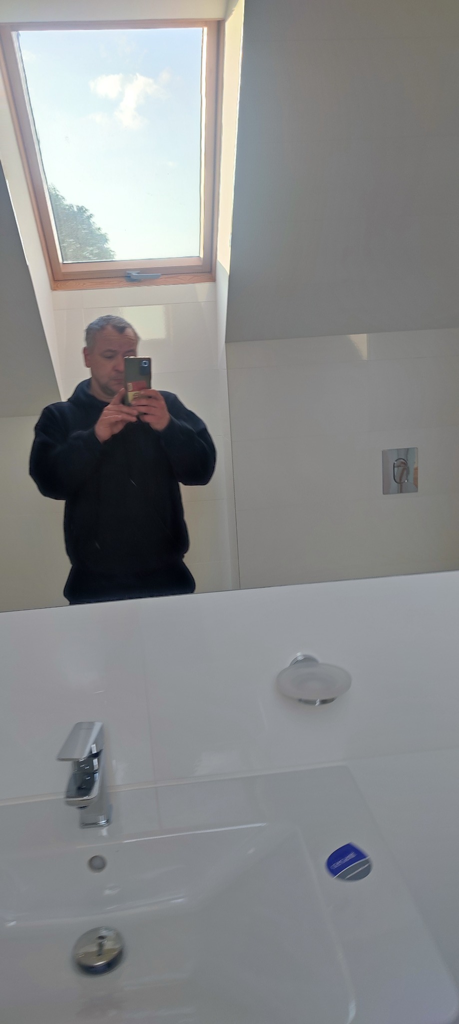 Mężczyzna robiący sobie zdjęcie w lustrze w nowocześnie wykończonej łazience z oknem dachowym, widoczna biała umywalka, bateria i dozownik na mydło.