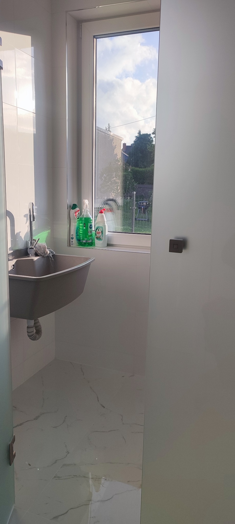 Nowoczesna łazienka z szarą umywalką pod oknem, białe płytki imitujące marmur na podłodze, środki czystości na parapecie.