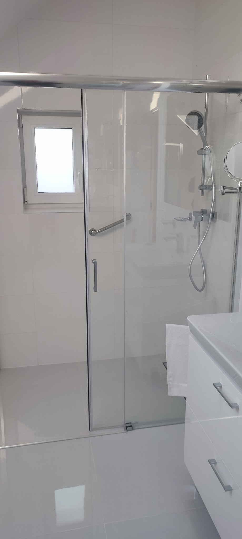 Nowoczesna, jasna łazienka z kabiną prysznicową ze szklanymi przesuwnymi drzwiami, chromowaną armaturą, umywalką z szafką z szufladami oraz małym oknem.