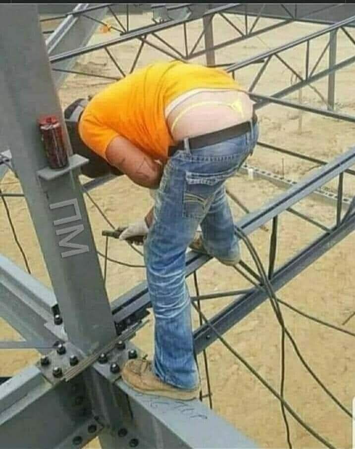 Spawacz w pomarańczowej koszulce i dżinsach spawa stalową konstrukcję na zewnątrz, widoczny fragment bielizny ponad spodniami.
