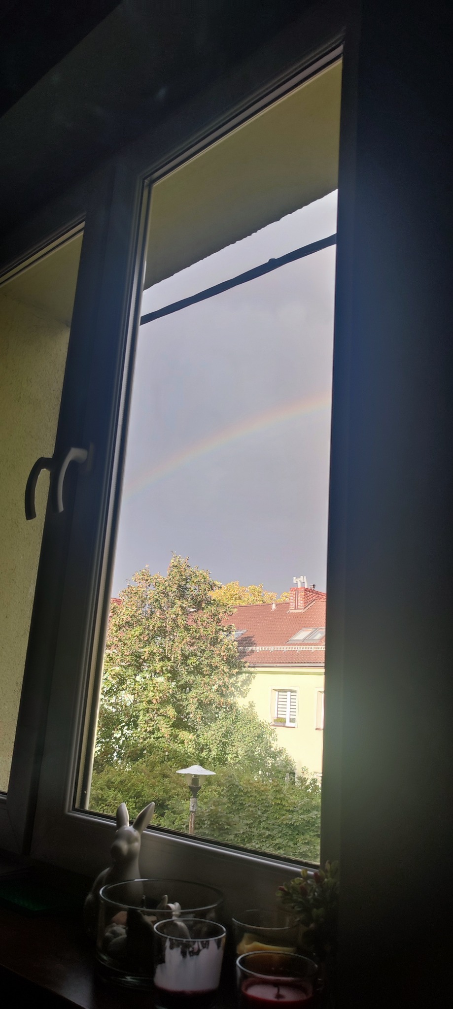 Widok z okna na tęczę nad budynkami i drzewami, z dekoracjami okiennymi w postaci szklanych naczyń i ceramicznego zajączka.