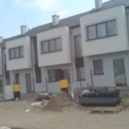 Szereg identycznych domów w zabudowie bliźniaczej z ciemnymi dachami, widoczne materiały budowlane i teren w trakcie prac wykończeniowych.