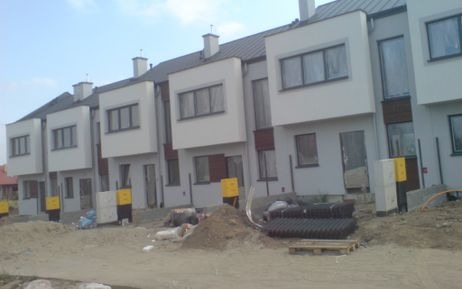 Szereg identycznych domów w zabudowie bliźniaczej z ciemnymi dachami, widoczne materiały budowlane i teren w trakcie prac wykończeniowych.