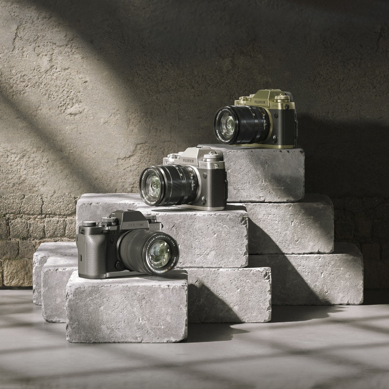 Trzy aparaty Fujifilm X-T3 w kolorach czarnym, srebrnym i oliwkowym, ustawione na stopniowanej konstrukcji z szarych betonowych bloków na tle teksturowanej ściany z cieniem.