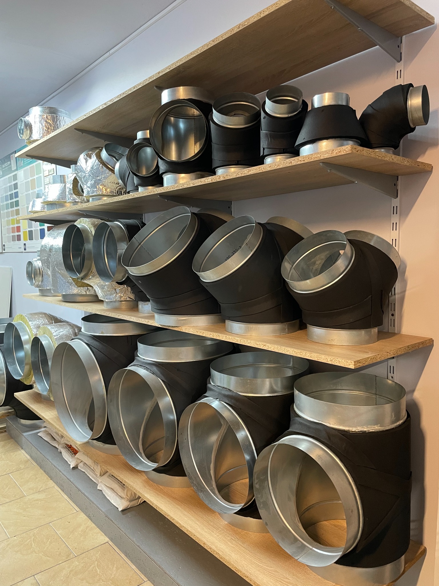 Różne kształtki wentylacyjne, niektóre zaizolowane, ułożone na drewnianych półkach w magazynie.