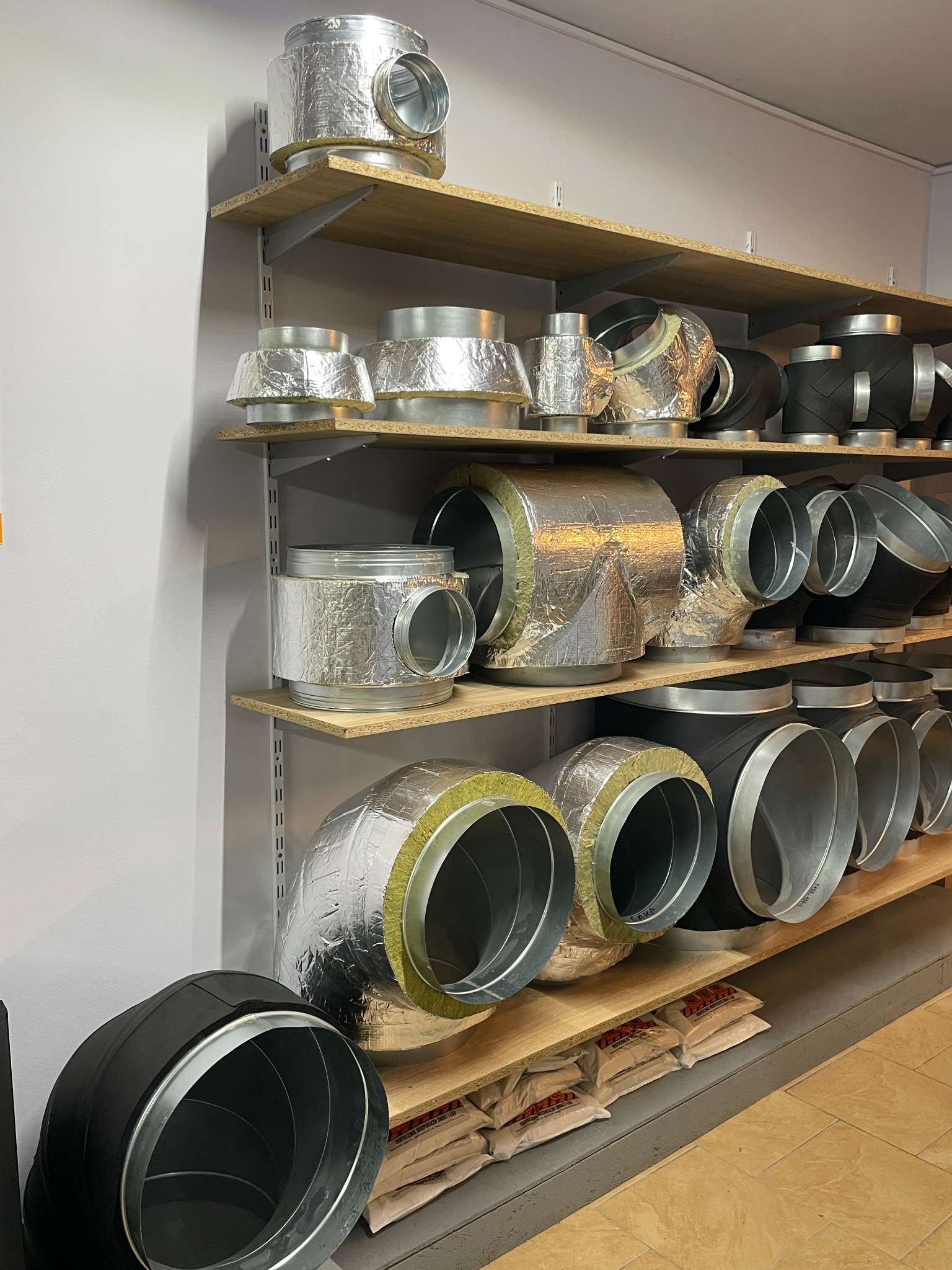 Półki w magazynie z różnymi kształtkami wentylacyjnymi, w tym kolanka i redukcje, niektóre z izolacją termiczną z wełny mineralnej i folii aluminiowej.