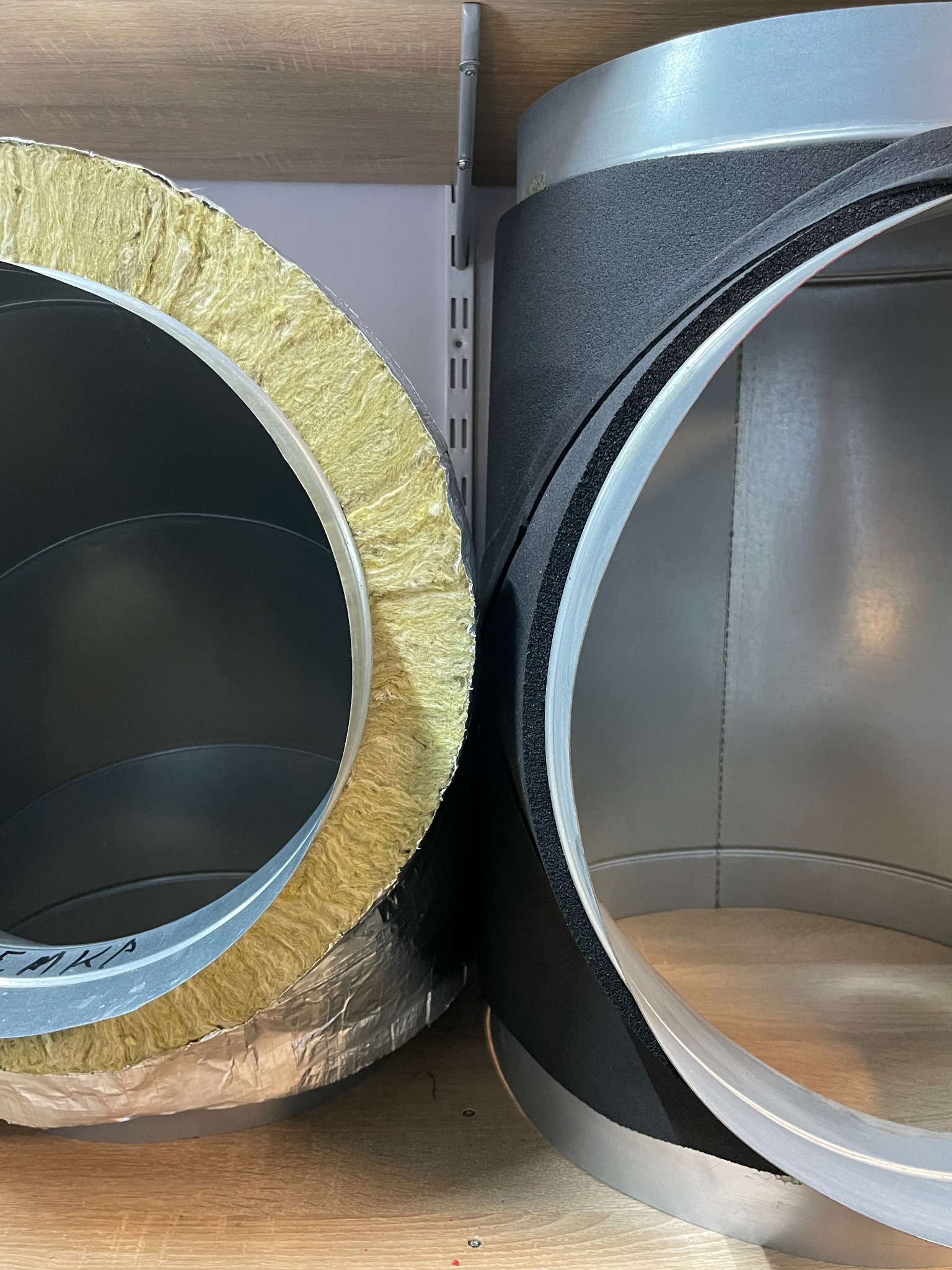 Dwa odcinki izolowanych kanałów wentylacyjnych: jeden z wełną mineralną i folią aluminiową, drugi z czarną pianką izolacyjną, widoczne od strony przekroju poprzecznego.