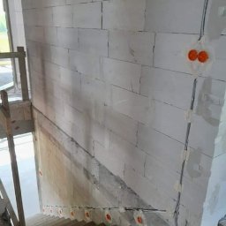 INSTAL-POL USŁUGI ELEKTRYCZNE - Wewnętrzne ściany z betonu komórkowego w trakcie budowy, widoczne puszki elektryczne i przewody prowadzone po ścianie w peszlu, schody w surowym stanie.