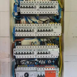 Rozdzielnia elektryczna z zainstalowanymi bezpiecznikami i przewodami o różnych kolorach, widok od frontu na ścianie z szarego bloczka.