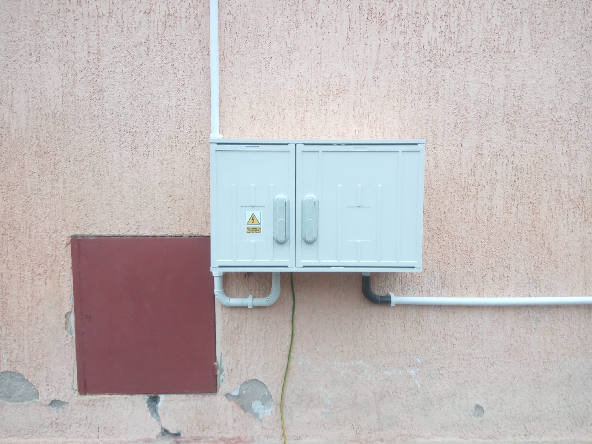 Zewnętrzna szafka elektryczna z podwójnymi drzwiczkami, zamontowana na ścianie o nierównej fakturze z widocznymi rurami PCV i uziemieniem.