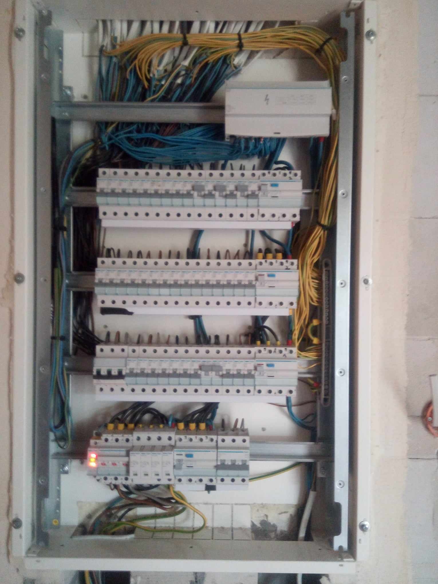 Otwarta skrzynka elektryczna z widocznymi bezpiecznikami, przewodami w kolorach niebieskim, żółtym i zielonym, z jednym włączonym bezpiecznikiem sygnalizowanym czerwoną diodą, zamontowana...