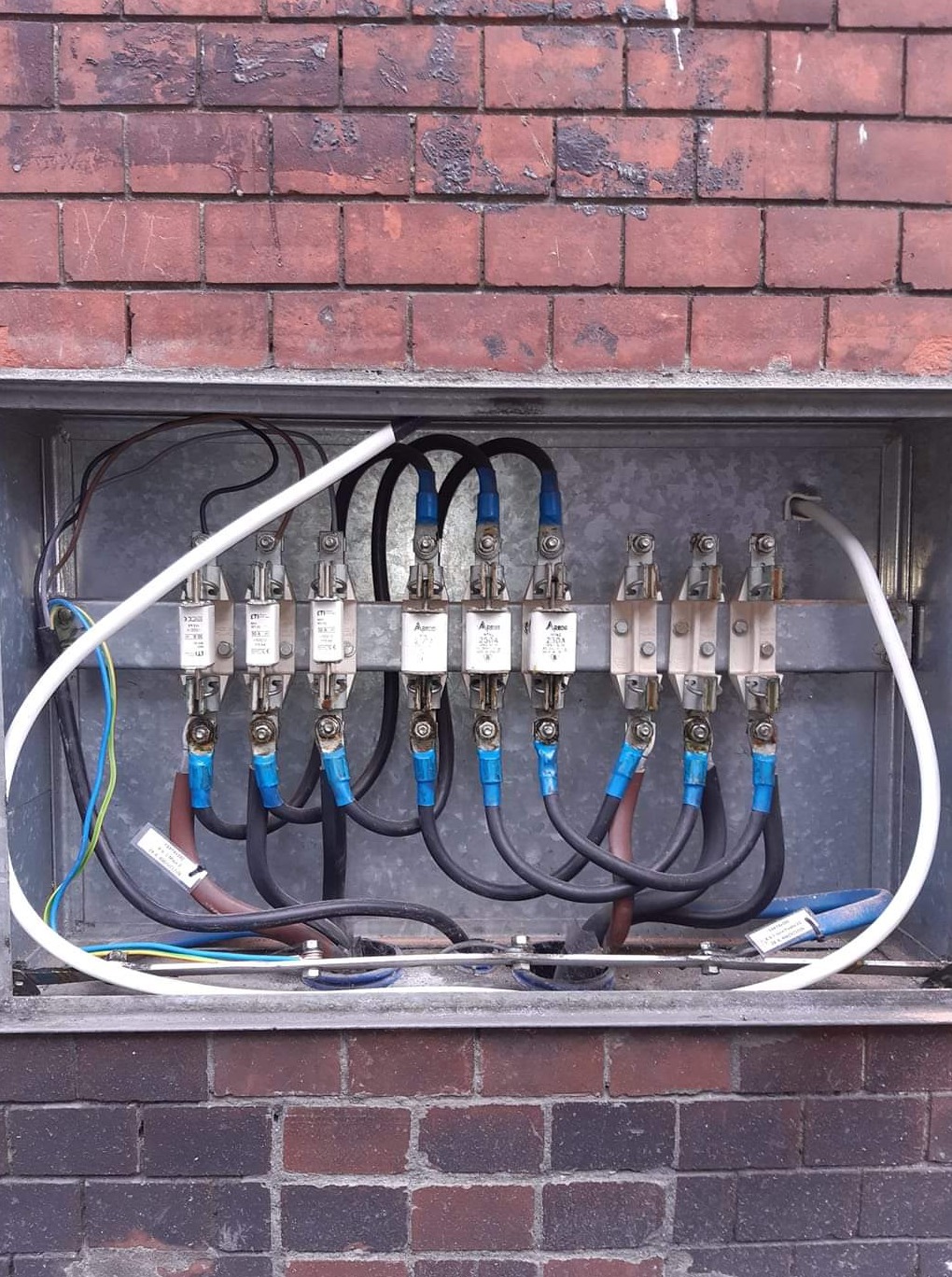 Otwarta skrzynka z bezpiecznikami na tle ceglanej ściany, widoczne kable w niebieskiej izolacji i białe rurki ochronne.
