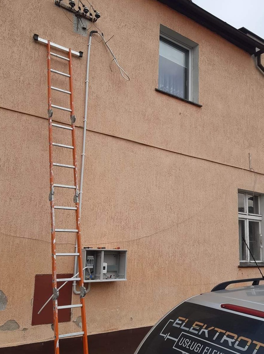 Montaż skrzynki elektrycznej na elewacji budynku w Świebodzinie, widoczna drabina oparta o ścianę i częściowo samochód z logo firmy elektrycznej.