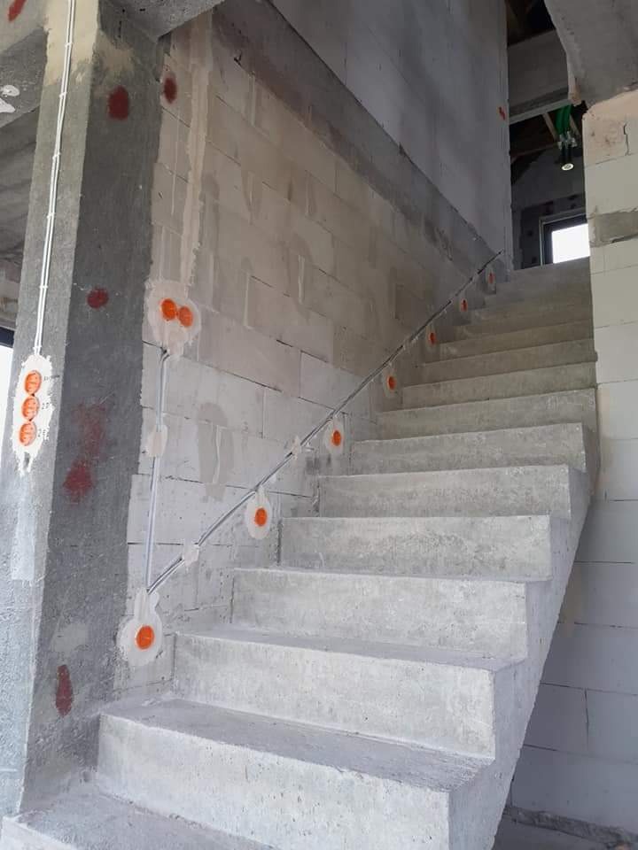 Surowe betonowe schody w trakcie budowy z widocznymi puszkami instalacyjnymi na ścianach i przewodami elektrycznymi prowadzonymi po powierzchni.