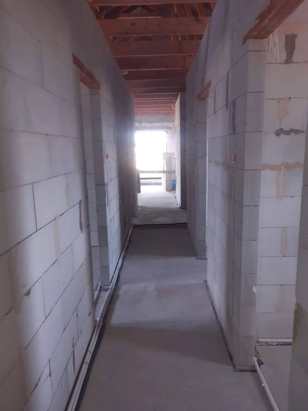 Wnętrze surowego budynku w trakcie budowy, widoczne ściany z bloczków, drewniany strop i rury instalacji elektrycznej prowadzone wzdłuż ścian.