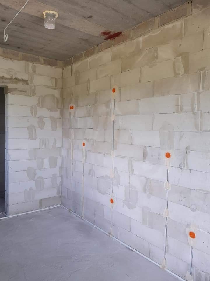 Wnętrze w stanie surowym z widocznymi przewodami elektrycznymi prowadzonymi po ścianach z betonu komórkowego, zabezpieczonymi pomarańczowymi puszkami i białym gipsem.