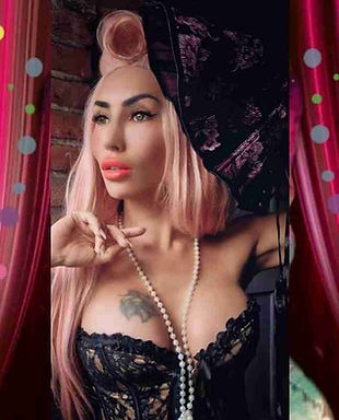 Kobieta z różowymi włosami, w czarnym gorsecie i perłami, pozuje na tle ceglanej ściany i dekoracji teatralnych, trzymając wachlarz. Wyrazisty makijaż.