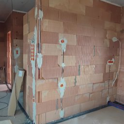 EL-craft Rafał Rajtar - Wnętrze w trakcie budowy z czerwonej cegły, widoczne kable elektryczne w bruzdach zalanych gipsem, otwory pod gniazdka i rury instalacji wodnej przy podłodze.