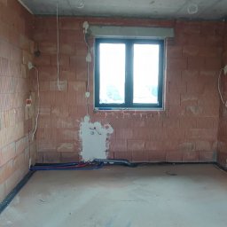 EL-craft Rafał Rajtar - Surowy stan wnętrza domu z widoczną instalacją elektryczną w trakcie montażu, ściany z czerwonej cegły, okno z ciemną ramą, kable i puszki elektryczne wystające ze ścian.