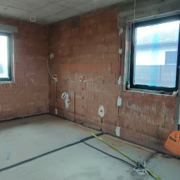 EL-craft Rafał Rajtar - Surowy stan wnętrza domu murowanego w trakcie budowy: widoczne instalacje elektryczne w ścianach z pustaków ceramicznych, okna z folią ochronną, prowizoryczne kable na podłodze i pomarańczowa...