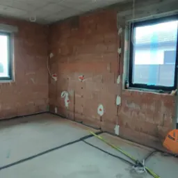 Surowy stan wnętrza domu murowanego w trakcie budowy: widoczne instalacje elektryczne w ścianach z pustaków ceramicznych, okna z folią ochronną, prowizoryczne kable na podłodze i pomarańczowa...