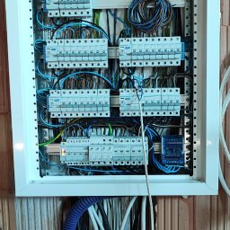 EL-craft Rafał Rajtar - Rozdzielnica elektryczna Zenergy zamontowana na ścianie z pustaków ceramicznych, z widocznymi zabezpieczeniami i przewodami w różnych kolorach, w tym niebieska rura karbowana doprowadzająca kable.